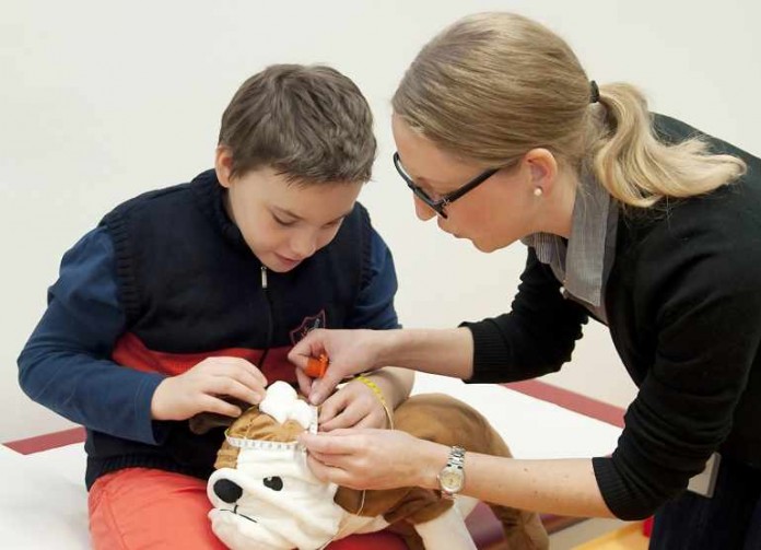Dr. Ann-Sophie Kaiser, Ärztin der neuropädiatrisch-genetischen Sprechstunde, zeigt Julian an einem Plüschtier, wie sie den Kopfumfang messen wird